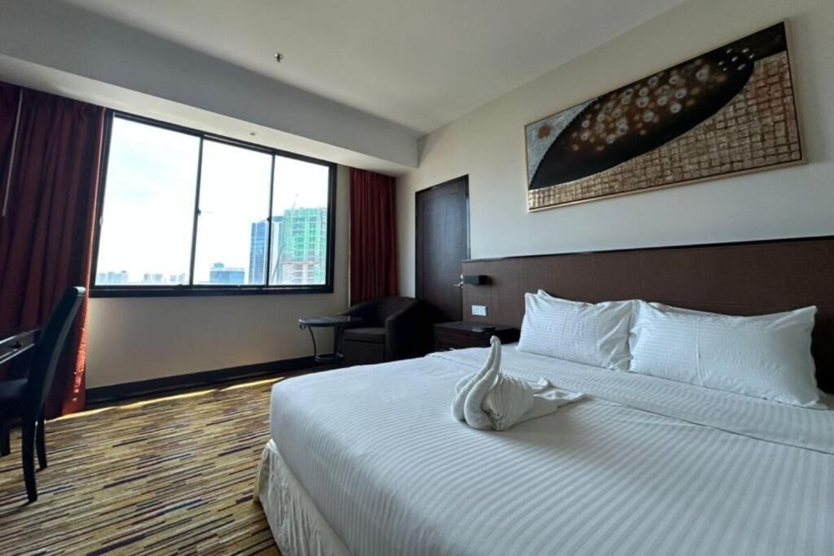 Habitación de hotel con una cama grande, un escritorio, una silla y vistas de la ciudad a través del ventanal.