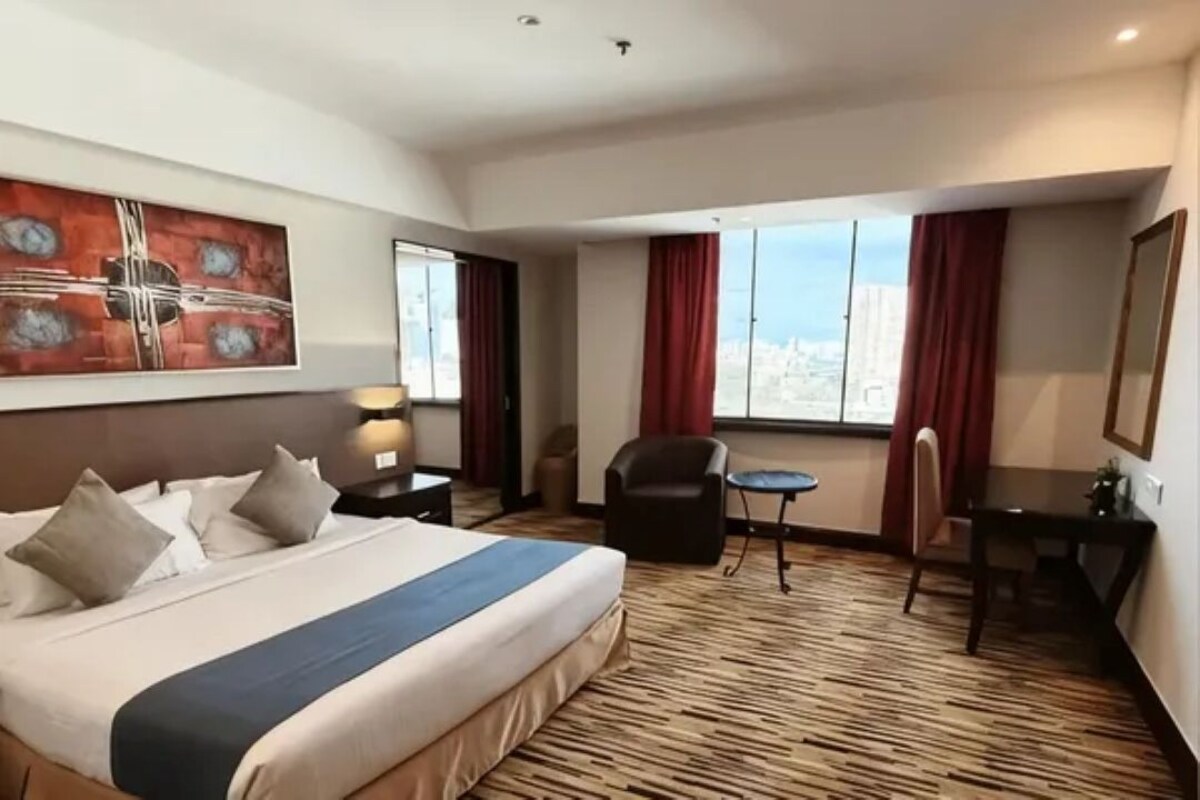 Habitación de hotel con una cama grande, un escritorio, una silla, una mesita y vistas a la ciudad por la ventana.
