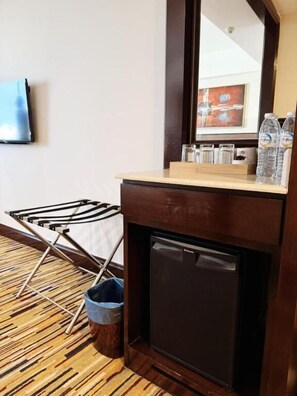 Una habitación con un armario de madera, un refrigerador pequeño, una silla plegable, un cesto de basura y un televisor fijado a la pared.