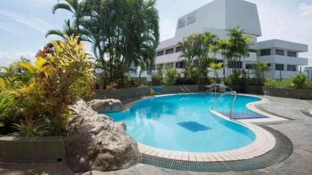 Una piscina con borde de baldosas, rodeada de plantas tropicales y un edificio al fondo.