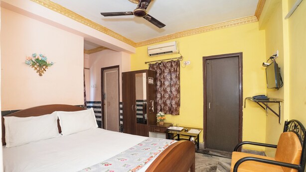 A hotel room with a bed, a desk, a chair, a ceiling fan, and a door. - Metro Stay Guest House (Barasat)