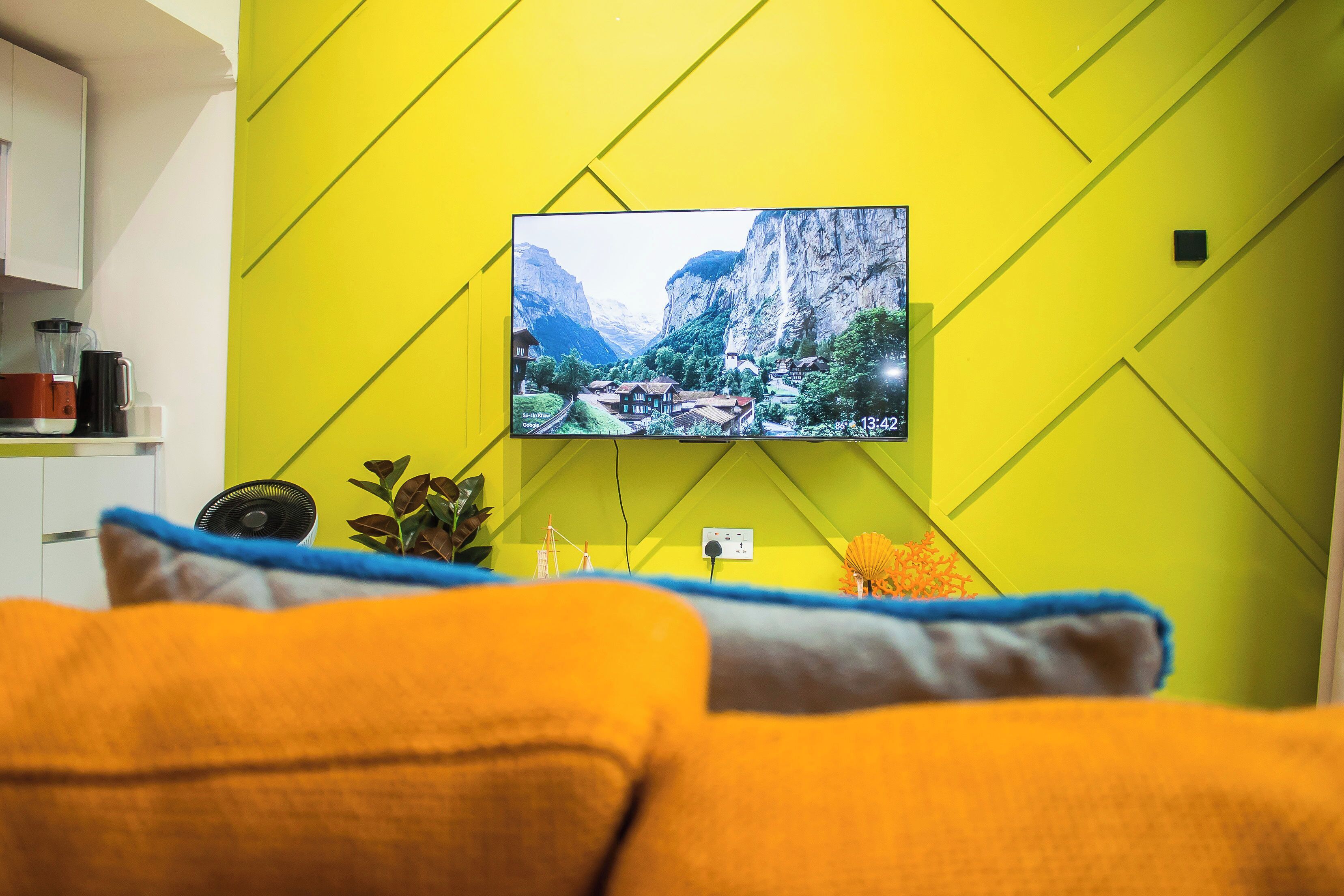 Una sala de estar con un televisor montado mostrando un paisaje montañoso, un sofá con cojines, un pequeño ventilador y una planta.