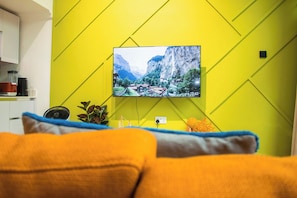 Una sala de estar con un televisor montado mostrando un paisaje montañoso, un sofá con cojines, un pequeño ventilador y una planta.
