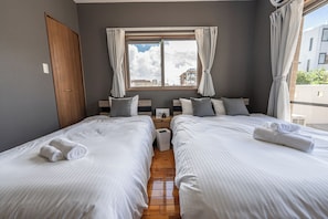 A hotel room with two beds, a window with curtains, a wooden door, and a view of buildings outside. - Komachi House (Chatan)