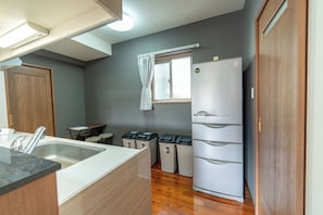 A compact kitchen with a sink, countertop, wooden cabinets, a refrigerator, and a window with curtains. - Komachi House (Chatan)