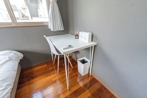 A small, neatly arranged bedroom with a single bed, a white desk with a chair, a small trash bin, and a window with a view of buildings. - Komachi House (Chatan)