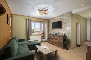 A living room with a green sofa, a wooden coffee table, a stone wall, a mounted TV, and a window with curtains.