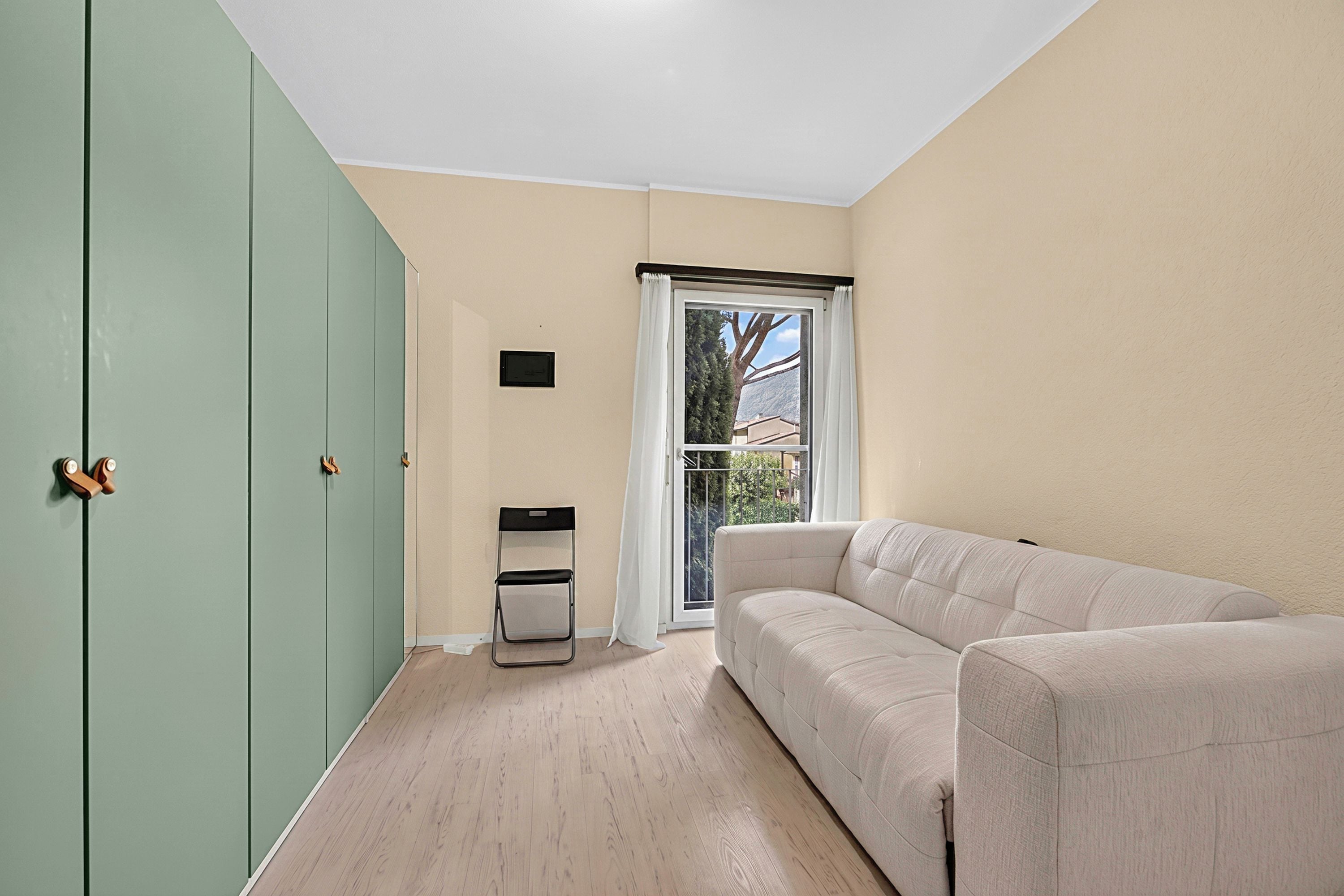 A room with a green wardrobe, a white sofa, a small table, and a window with a view of a tree and buildings.