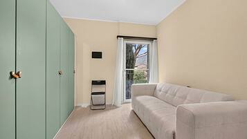 Une pièce avec une armoire verte, un canapé blanc, une petite table et une fenêtre donnant sur un arbre et des immeubles.