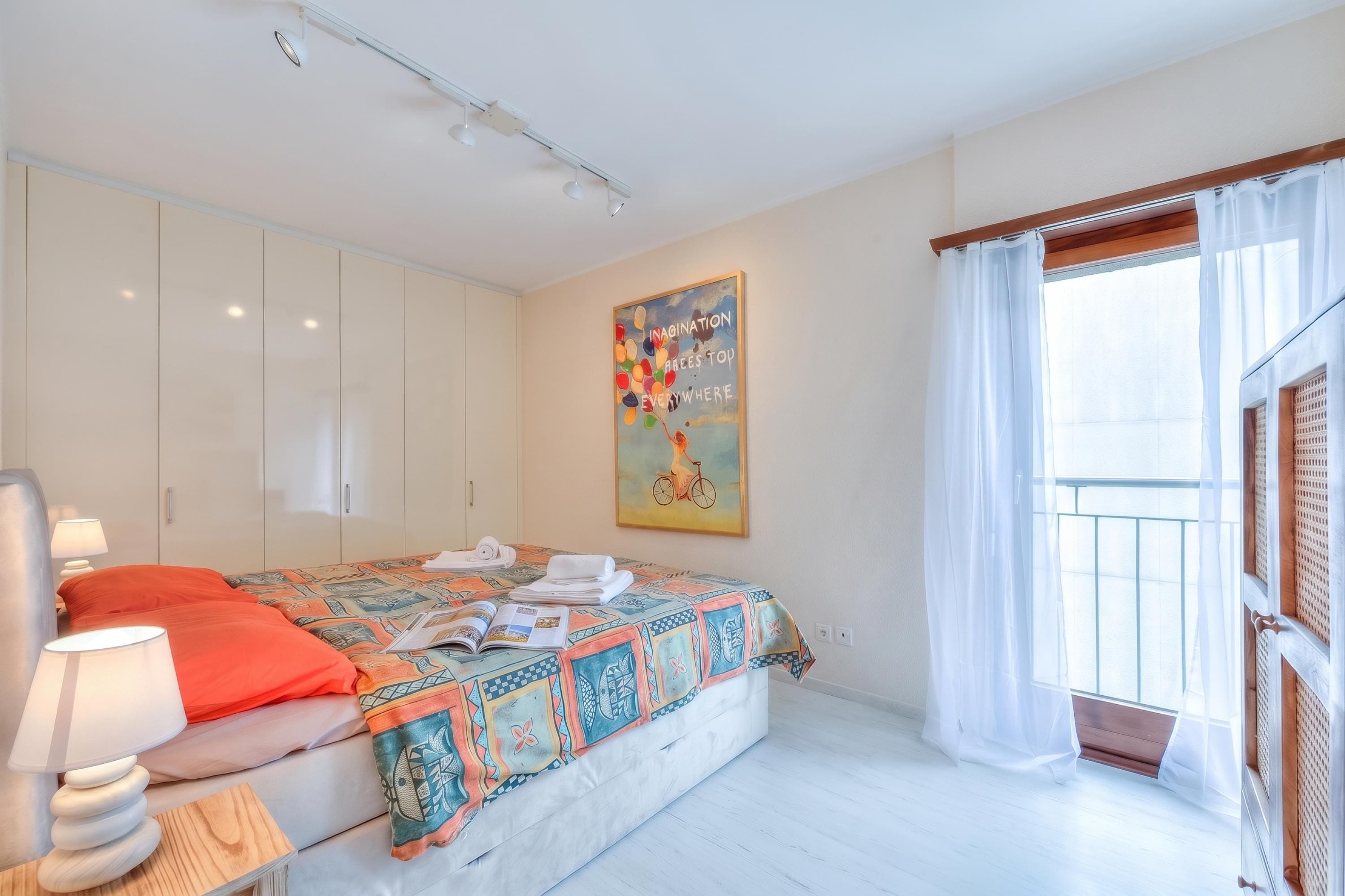 A bedroom with a bed, a nightstand, a lamp, a painting, and a window with curtains.