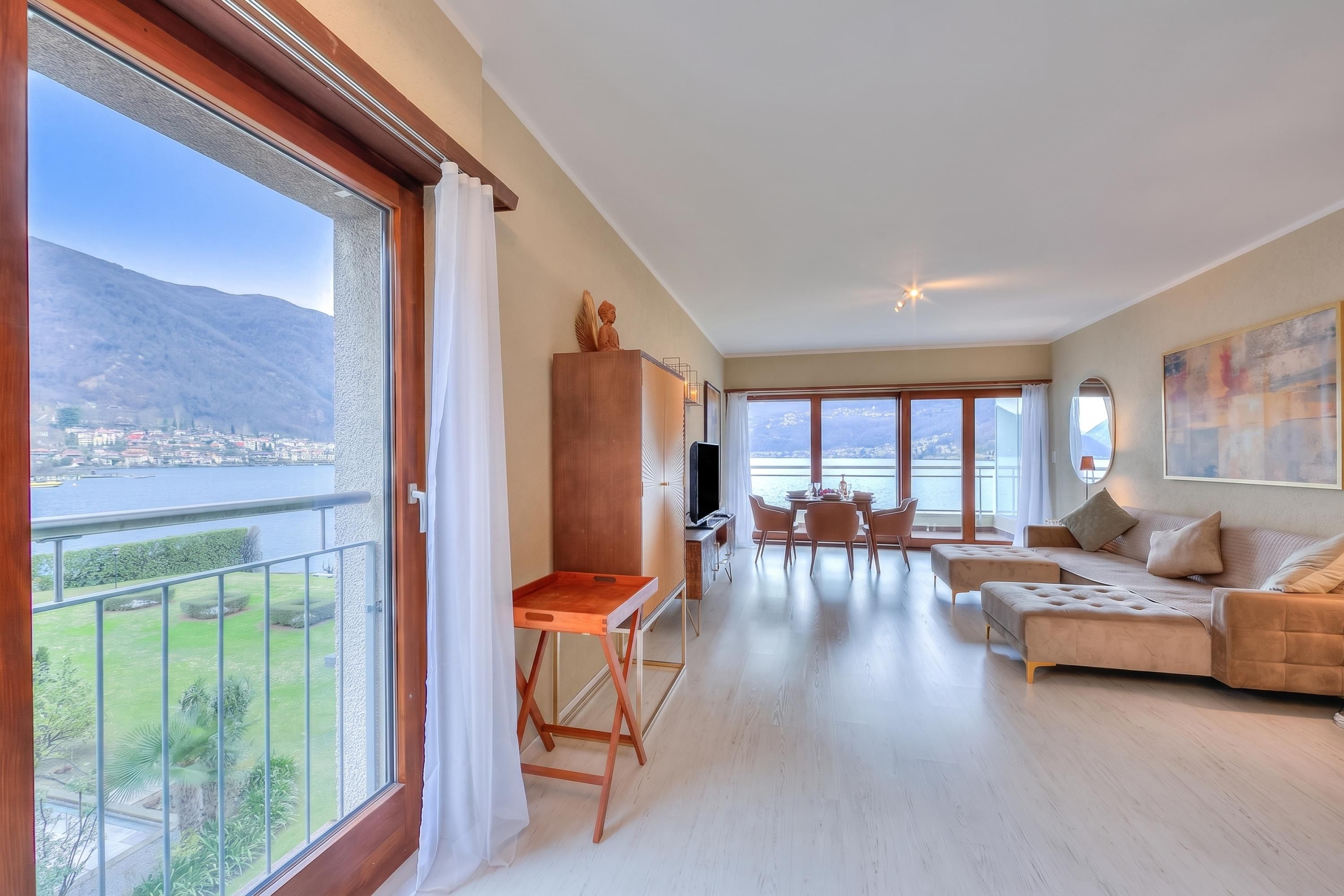A spacious living area with a balcony overlooking a lake and mountains, a wooden cabinet, a television, and a sofa.