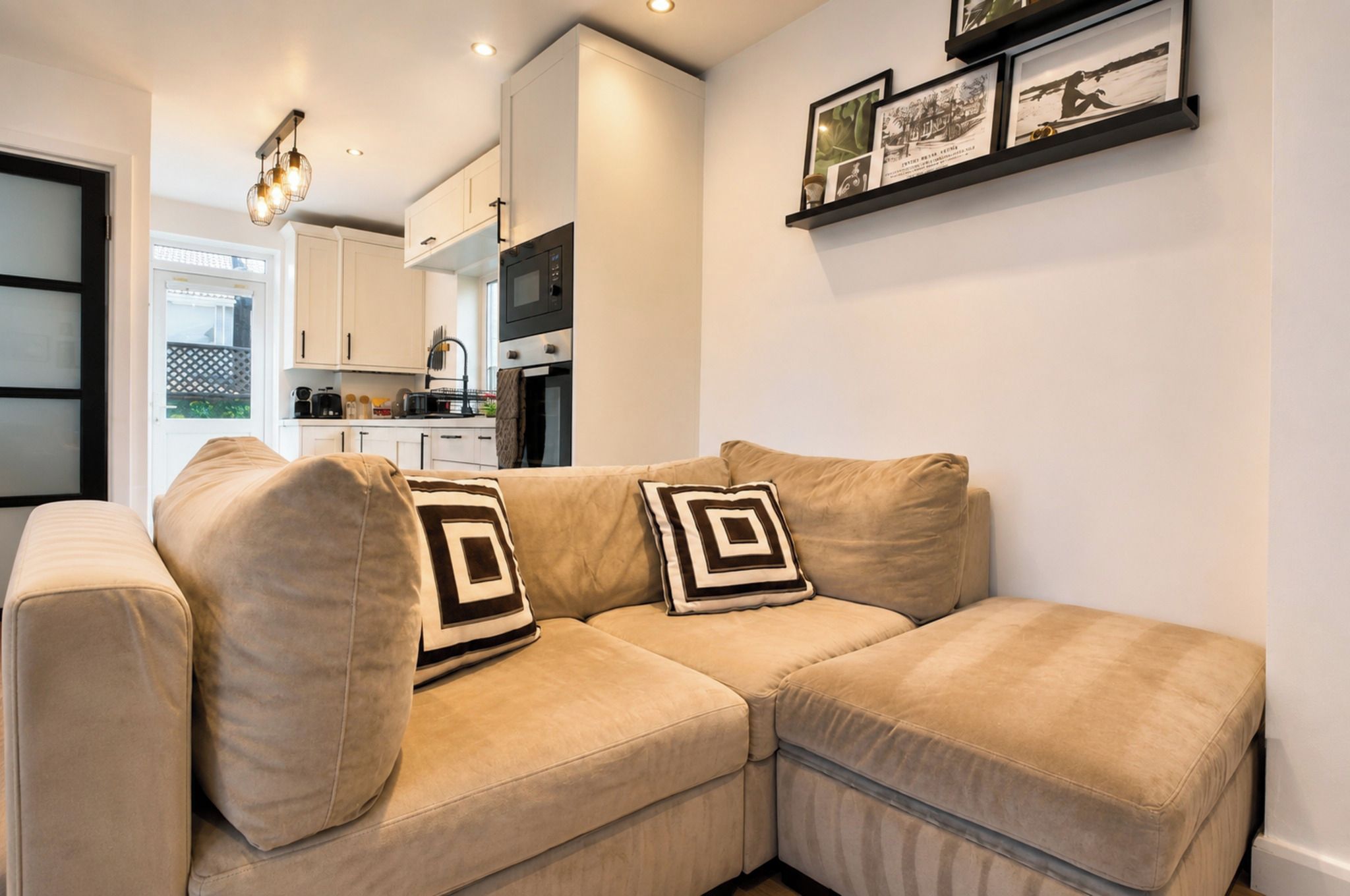 Una sala de estar moderna con un sofá seccional color beige, una cocina al fondo y una pared con cuadros.