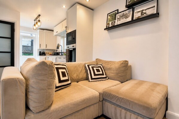 A modern living room with a beige sectional sofa, a kitchen in the background, and a wall with framed pictures.