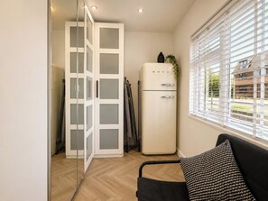 Una habitación compacta con puerta de vidrio corredera, un refrigerador blanco, un sofá negro con cojines estampados y una ventana con persianas.