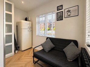 A compact living space with a kitchenette, a sofa with patterned cushions, a window with blinds, and framed artwork on the wall.