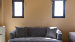 A living room with a grey sofa, two windows with blinds, and a white appliance on the left. - Kouvaras Equestrian Club Residence by RHS (Kouvara)