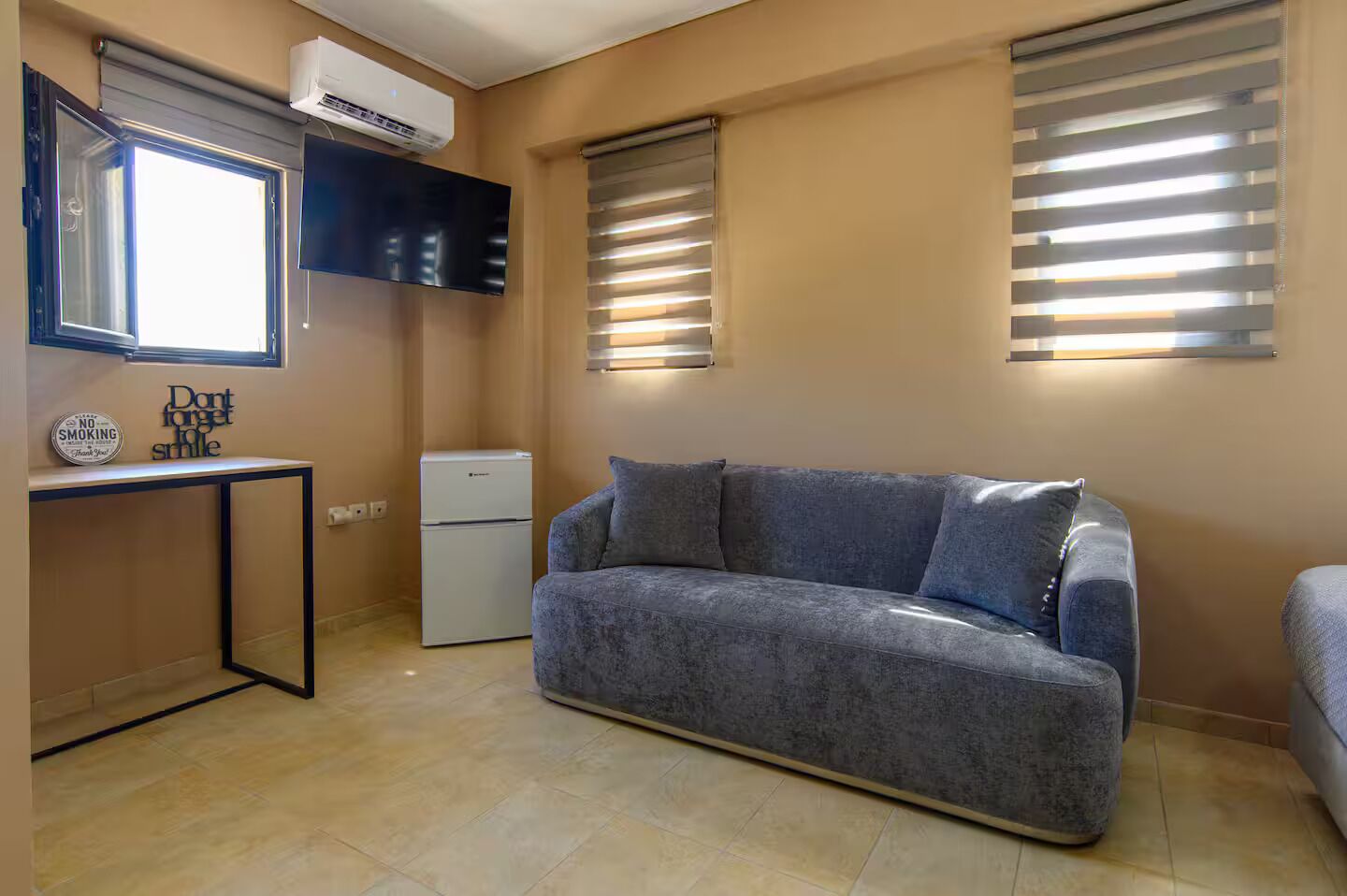 Un espace de vie compact comprenant un canapé, une petite kitchenette, un téléviseur à écran plat et une fenêtre avec des stores.