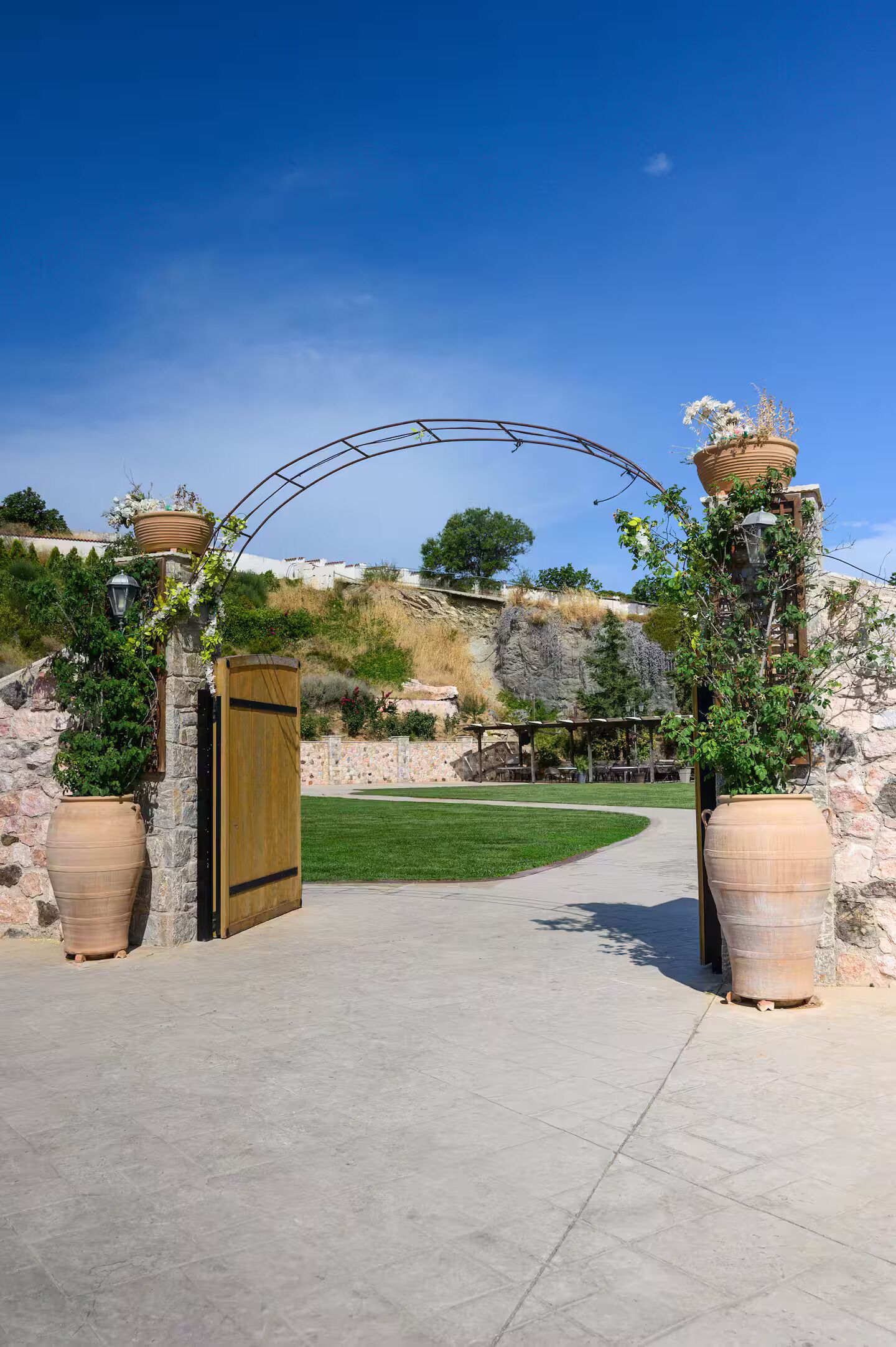 A paved pathway leading to a wooden gate, flanked by large terracotta pots with plants, under a clear blue sky.