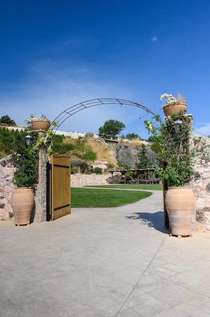 Un camino pavimentado que conduce a una puerta de madera, bordeado por macetas grandes de terracota con plantas, bajo un cielo azul despejado.