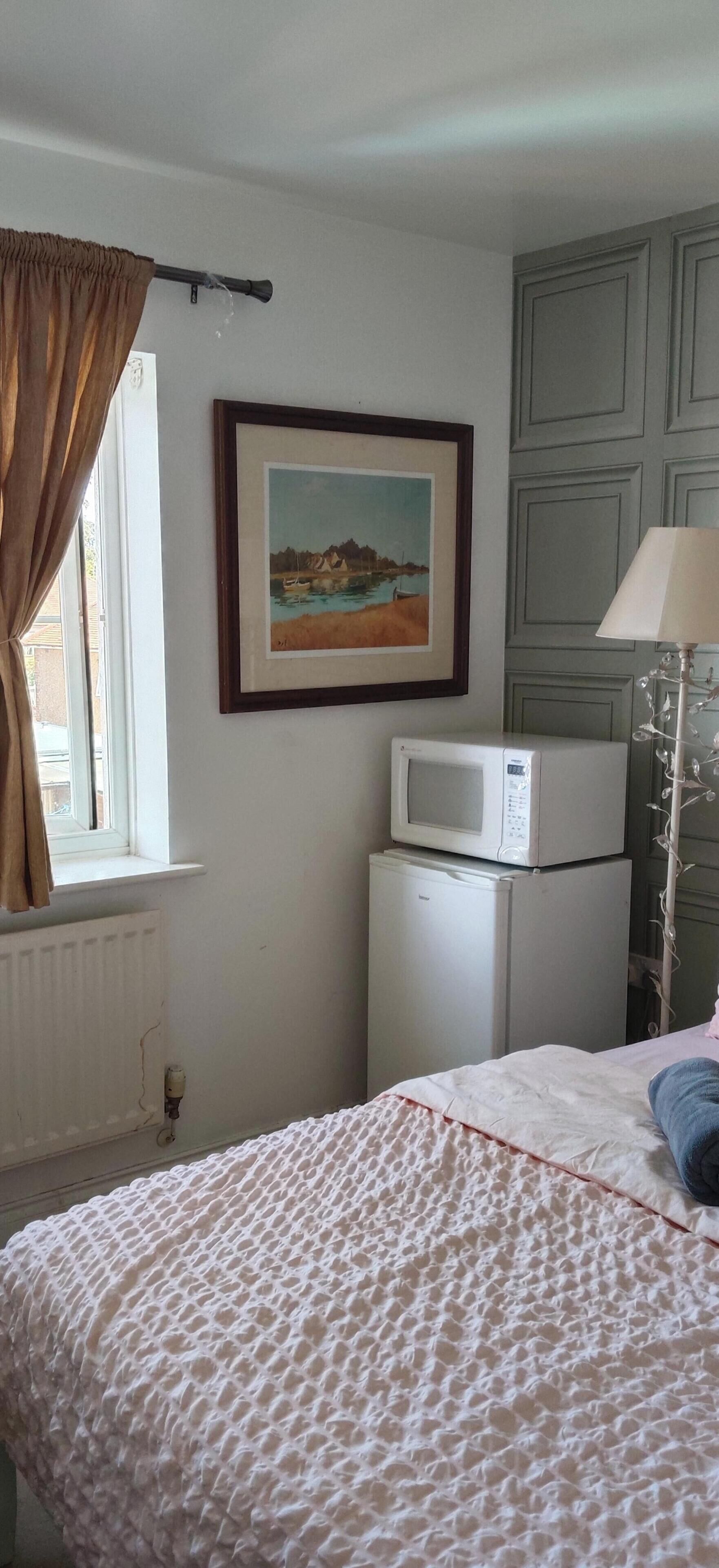 Ein Schlafzimmer mit einem Bett, einem Fenster mit Vorhängen, einem gerahmten Gemälde, einer Mikrowelle und einer Heizung.