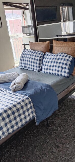 A bed with checkered bedding, a rolled white towel, and a view of the outside through a window.