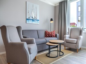 A modern living room with a grey sofa, two armchairs, a small wooden coffee table, and a painting of sailboats on the wall. - Balkonwohnung mit Blick ins Grune (Lübeck)
