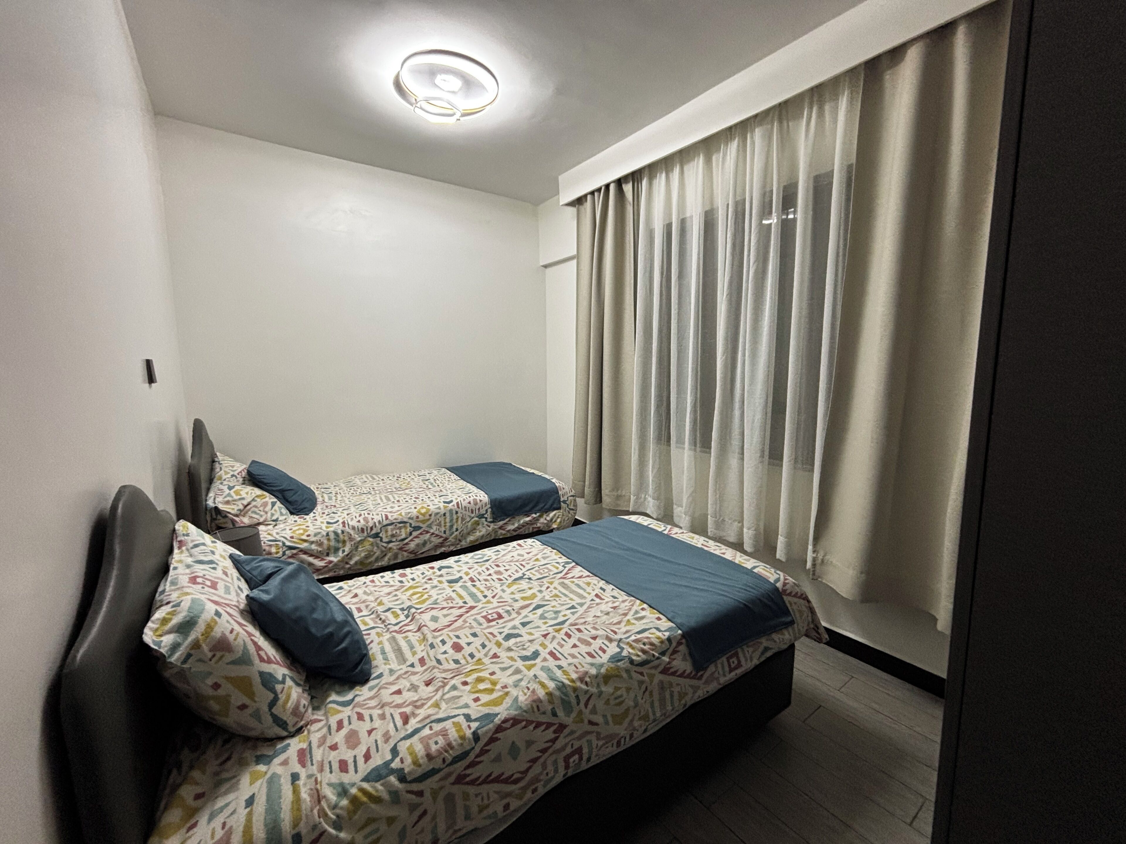 Une petite chambre moderne, dotée de deux lits, d’un luminaire au plafond et d’une fenêtre avec des rideaux.