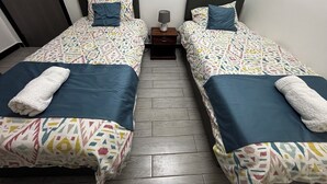 A hotel room with two beds, each with a patterned bedspread and pillows, a bedside table with a lamp, and a door to the left. - Takdiri Homes Neptune (Nairobi)