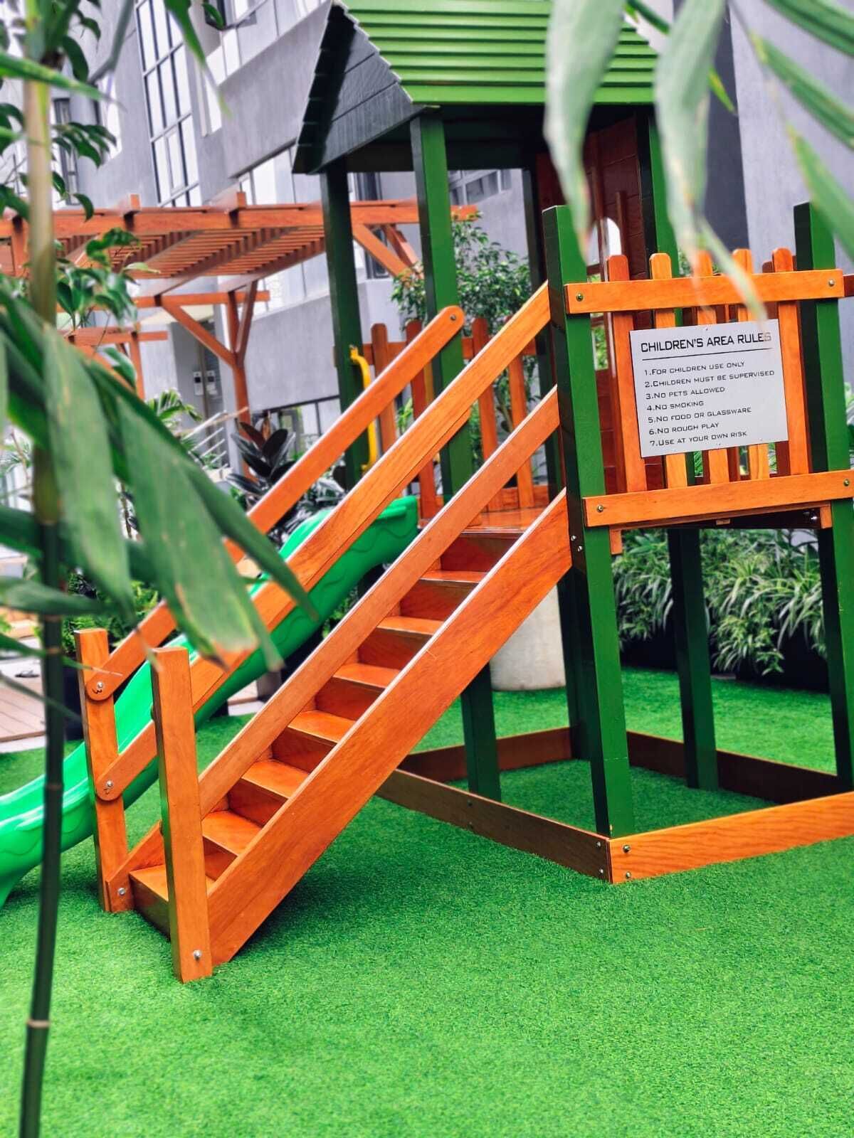 Un espace de jeux pour enfants avec un toboggan et des escaliers en bois, entouré de verdure et avec un bâtiment en arrière-plan.
