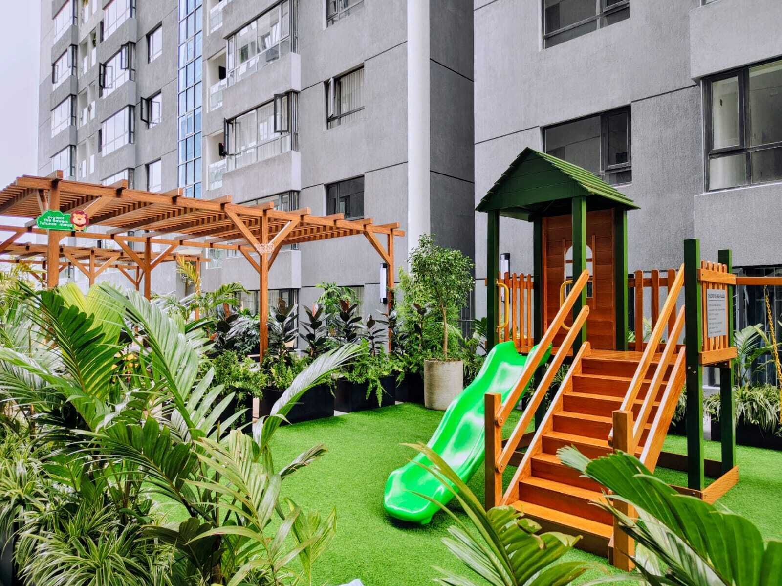 A children's play area with a slide and wooden play structures, surrounded by lush greenery.