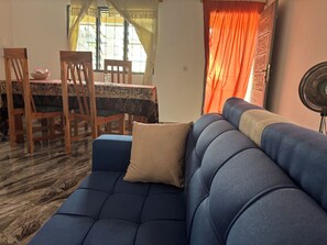 A living room with a blue sofa, wooden chairs, a dining table with a patterned tablecloth, and a fan. - Single-story house with an inner courtyard, ideal for a family (Abidjan)