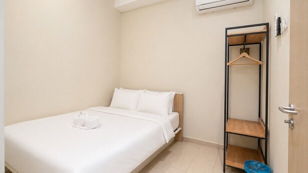 Habitación individual con una cama, cabecera de madera, cubrecama blanco, perchero y un cubo de basura azul.