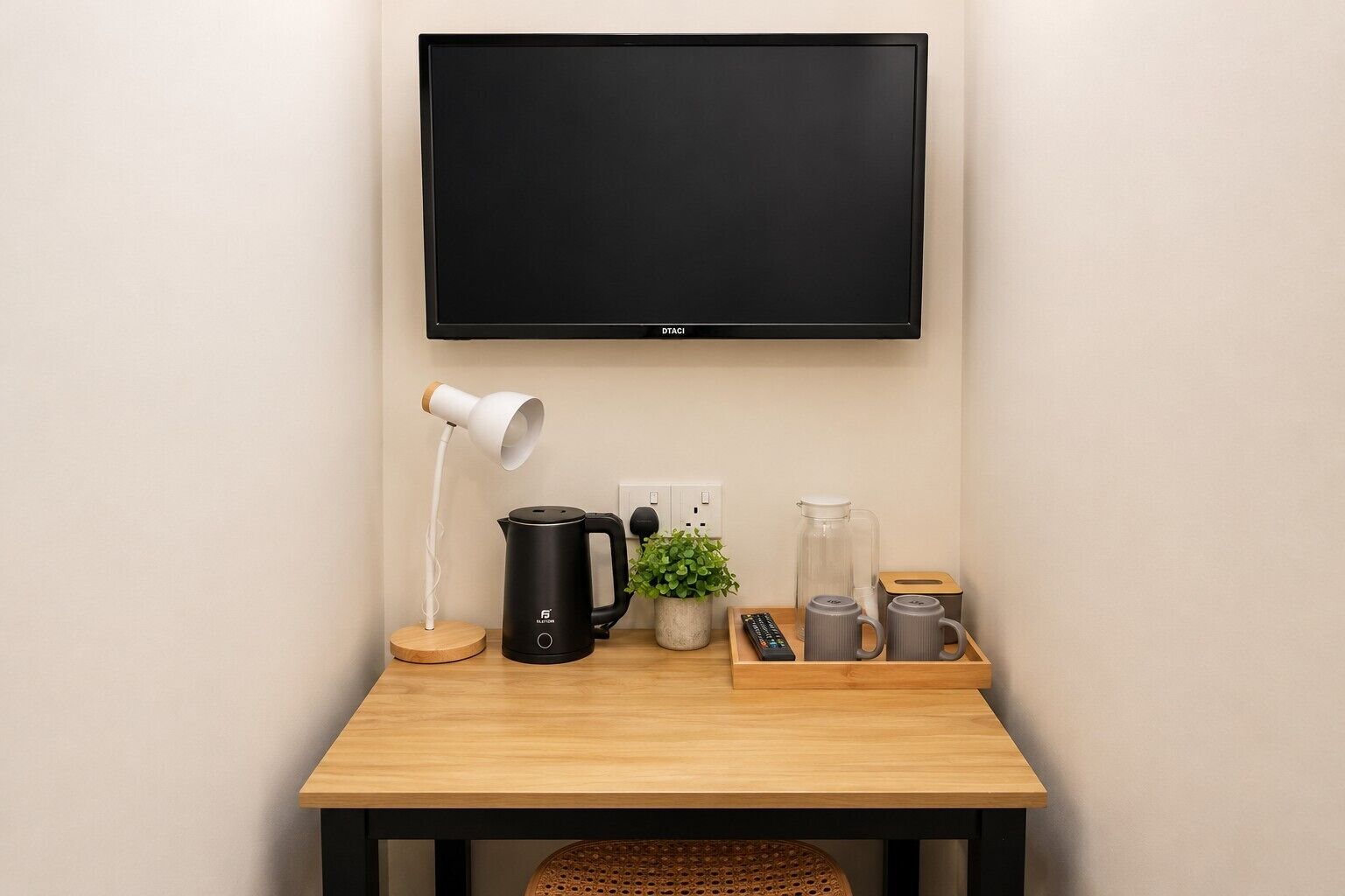 A room with a flat-screen TV mounted on the wall, a desk with a kettle, a coffee maker, a remote control, and a tray with mugs.