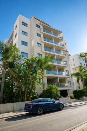 Un immeuble à plusieurs étages doté de balcons, une voiture garée devant et des plantes tropicales.