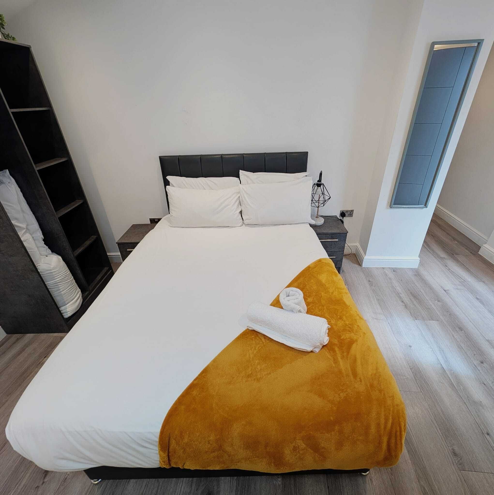 Un lit avec une literie blanche et un plaid de couleur moutarde, une table de chevet avec une lampe et une armoire avec un miroir.
