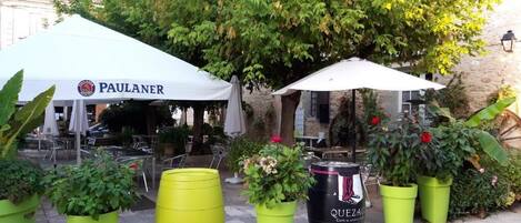 Mesa al aire libre con una carpa PAULANER y cartel de QUEZAC café, rodeado de plantas en macetas y una pared de piedra.