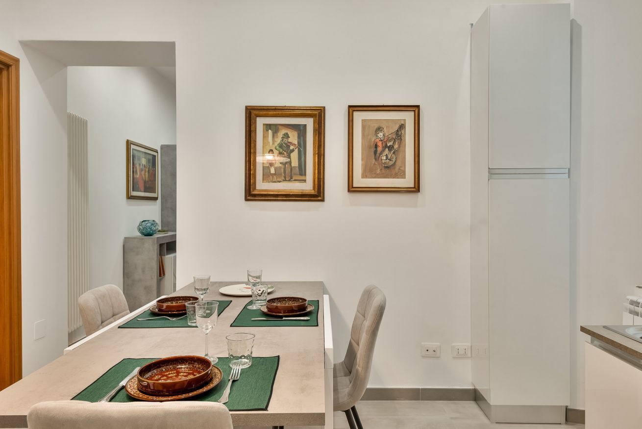 A dining area with a table set for two, framed artwork on the wall, and a modern kitchen with a white refrigerator.