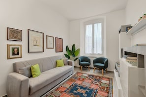 Un salón con sofá, dos sillones, alfombra estampada, cuadros en la pared y ventana con cortinas.
