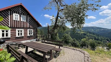 A wooden cabin with a picnic table and benches, surrounded by a gravel area and a view of a forested valley.