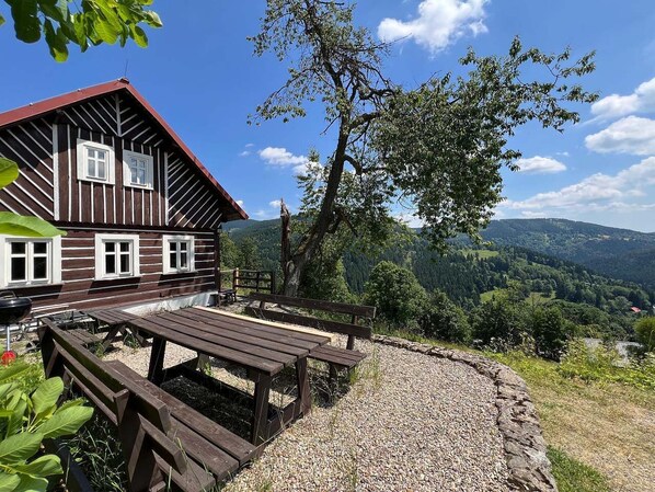 A wooden cabin with a picnic table and benches, surrounded by a gravel area and a view of a forested valley.