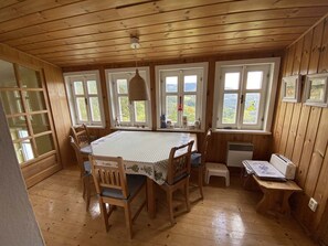 A wooden-paneled room with a dining table, chairs, a small stool, a radiator, and a window with a view of greenery.