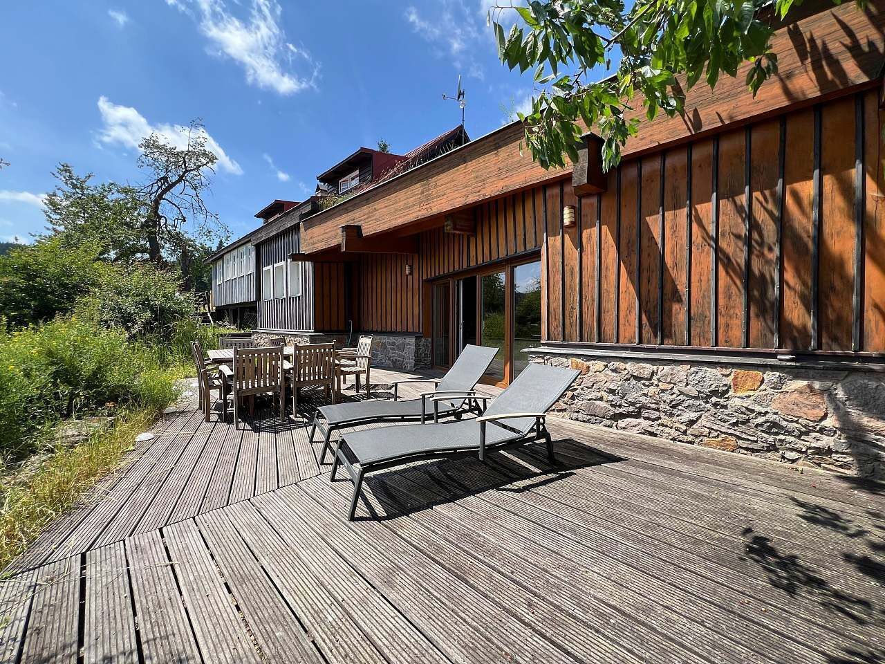 Une terrasse en bois avec des meubles d’extérieur, un mur en pierre et un bâtiment dont le revêtement extérieur est constitué de planches de bois disposées verticalement.