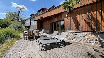 Une terrasse en bois avec des meubles d’extérieur, un mur en pierre et un bâtiment dont le revêtement extérieur est constitué de planches de bois disposées verticalement.