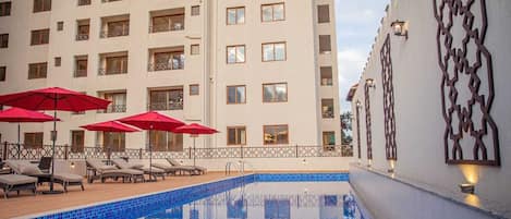 A hotel pool area with lounge chairs and red umbrellas, surrounded by a tall building with balconies.