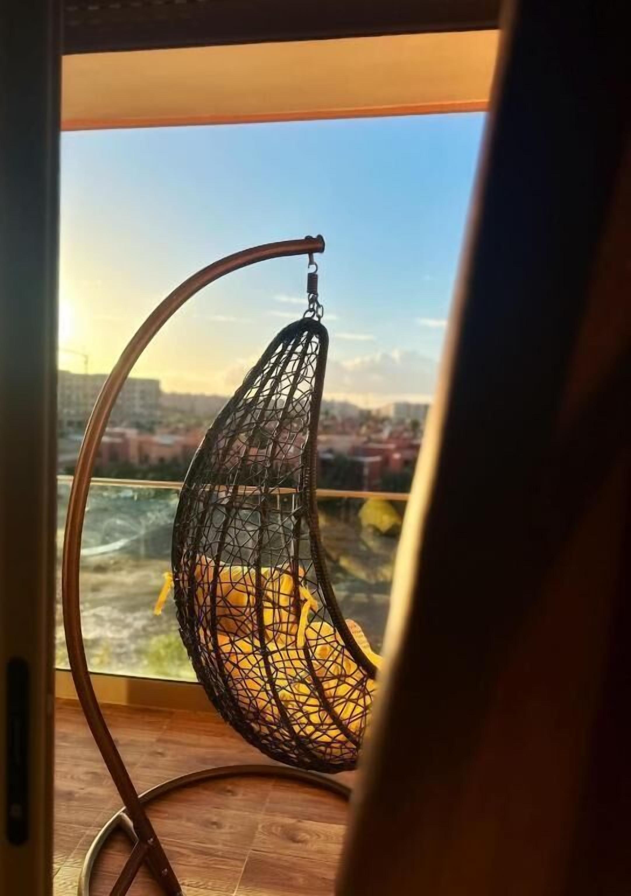 A hanging chair with a netted seat and a curved metal frame, positioned by a window overlooking a cityscape.