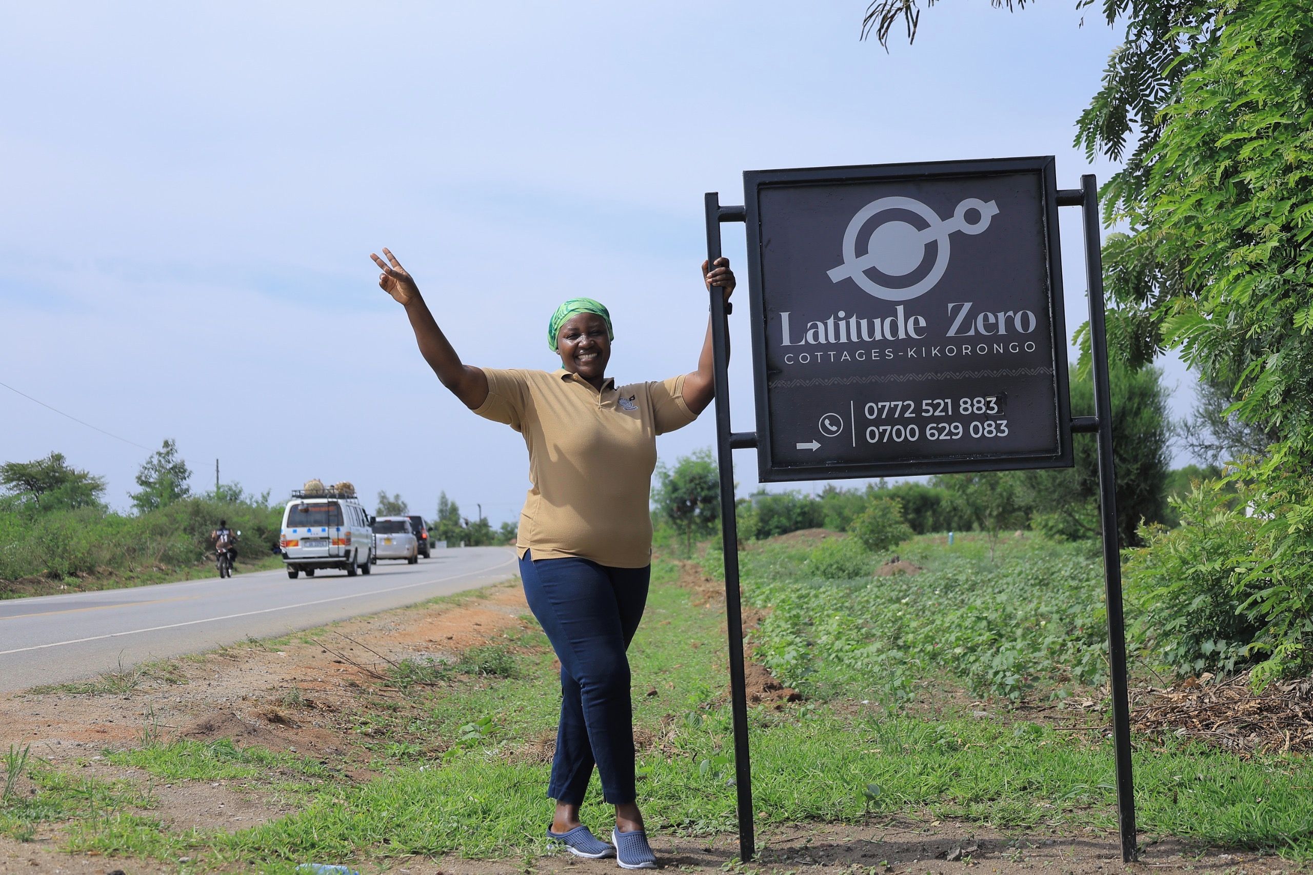 Eine Person steht neben einem Schild für Latitude Zero Cottages-Kikorongo mit Kontaktdaten.