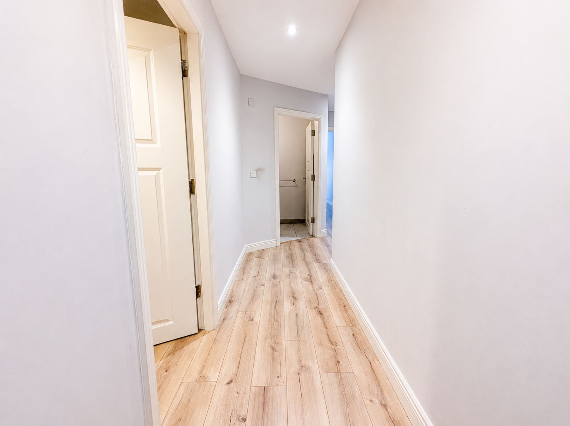 Un pasillo con suelo de madera y paredes blancas, que conduce a una puerta y un baño.