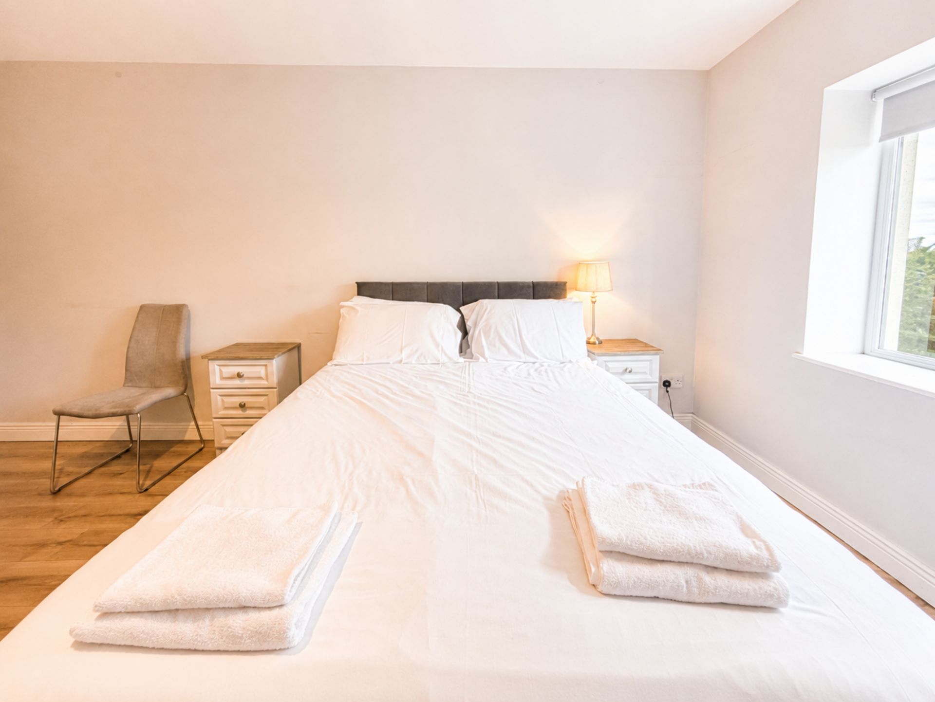A single bed with white linens, a bedside table with a lamp, a chair, and a window.