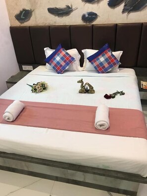 Ein ordentlich gemachtes Bett mit weißer und rosa Bettdecke, zwei Zierkissen, eine kleine Blumenkomposition und ein aufgerolltes Handtuch.