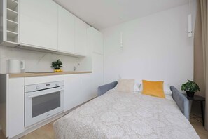 A modern bedroom with a bed, a small bedside table, a chair, a kitchenette with a stove, and a potted plant. - Functional Studio near Pole Mokotowskie Workspace Smart TV (Warszawa)